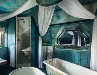 A bathroom with textured blue green walls and ceilings has hanging white draped curtains twin tubs and twin vanity