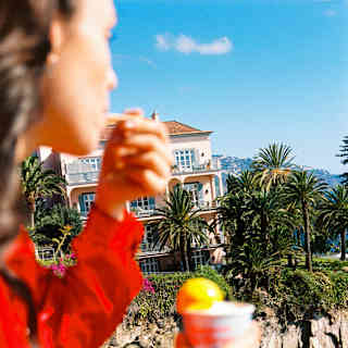 In soft-focus foreground, a woman in a red shirt enjoys a pot of orange-hued gelato, with Reid's Palace in crisp light behind.